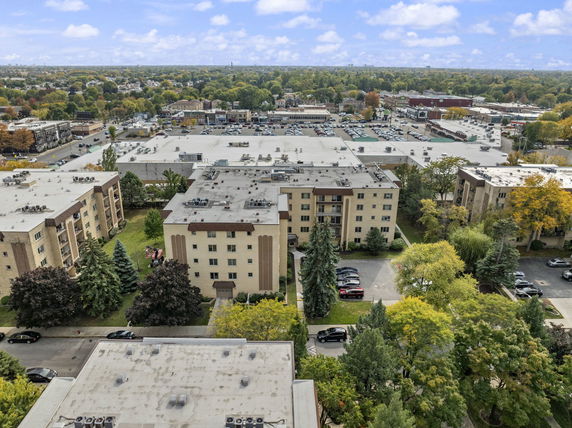 Aerial view of multiple apartment buildings with rooftops and parking areas, surrounded by trees and a suburban landscape.