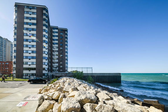 View of a tall residential building near a waterfront with rocks and water in the foreground.