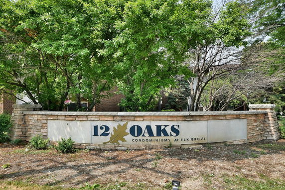 Front view of sign for 12 Oaks Condominiums with stone wall and surrounding trees.