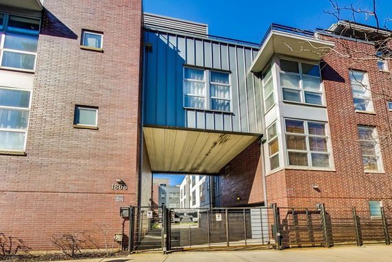 Front view of a modern multi-story building with brick and metal facade, featuring large windows and a gated entrance.