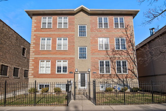 Front view of a multi-story brick apartment building with a central entrance and gated front yard.