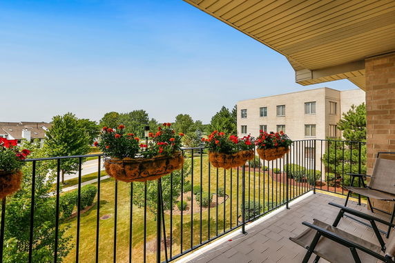 View from a balcony with hanging flower pots overlooking a landscaped area and neighboring buildings.