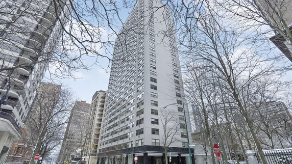 Front view of a tall, multi-story residential building with numerous windows and a street view with surrounding trees.