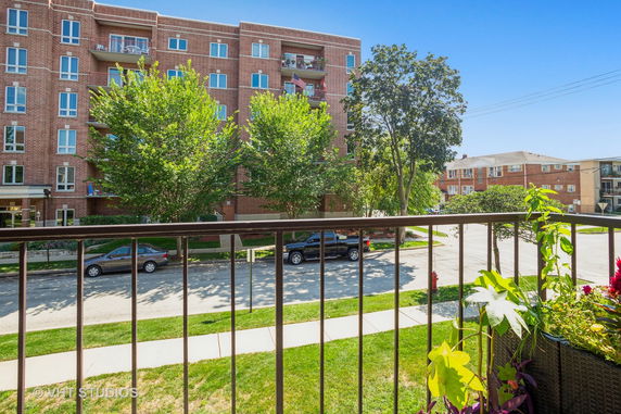 View from a balcony overlooking a street with parked cars and a multi-story brick building.