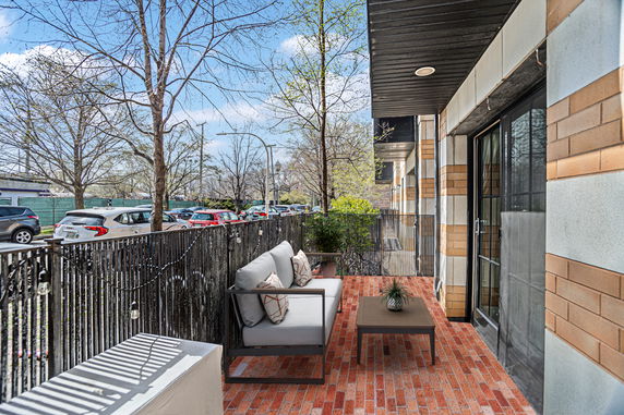 View of a patio area with seating next to a building, overlooking a street with parked cars and trees.