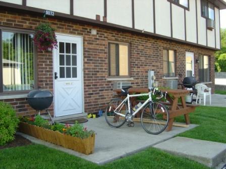 Front view of a brick exterior building with white door and windows, featuring a bicycle and patio items.