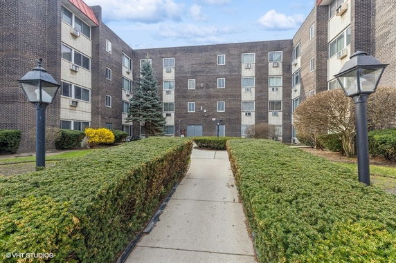 Front view of a multi-story brick apartment building with surrounding pathway and greenery.