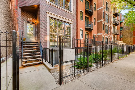 Front view of a multi-story brick building with stairs leading to the entrance and metal fencing.