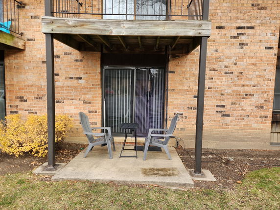 View of a building's exterior wall with a small patio area, featuring two chairs and a table under a balcony.