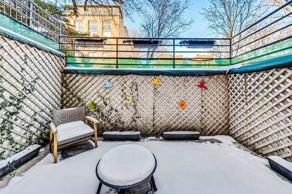 Outdoor patio area with snow-covered furniture and lattice walls, with a view of surrounding buildings.