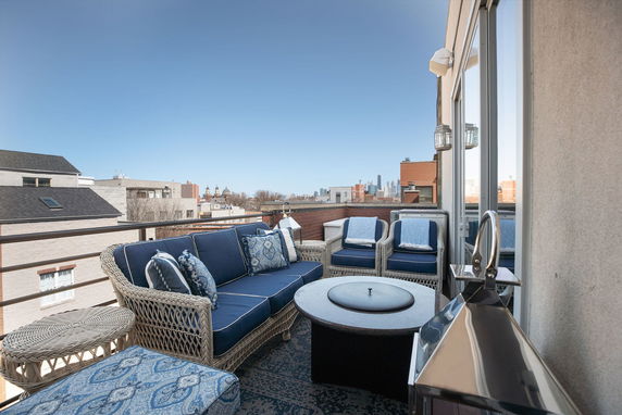 Rooftop seating area with wicker furniture and cushions, overlooking a cityscape.