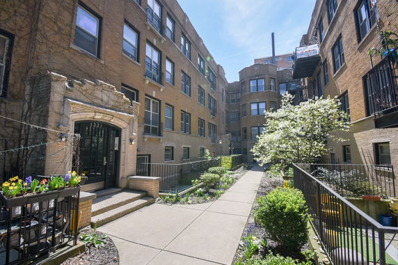 Front view of a multi-story brick apartment building with entrance and garden walkway.