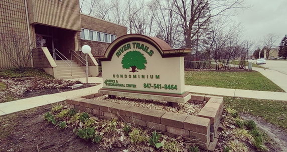 Front view of a condominium building with a sign labeled 'River Trails Condominium' and entrance steps.