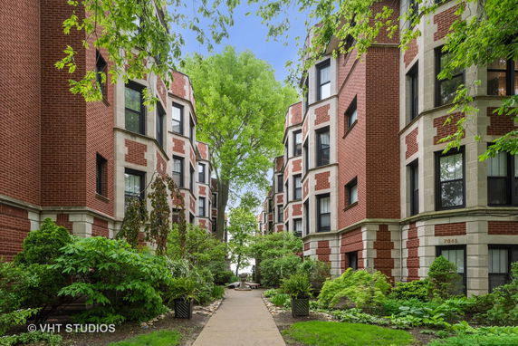 View of two brick apartment buildings with bay windows, separated by a pathway and surrounded by greenery.