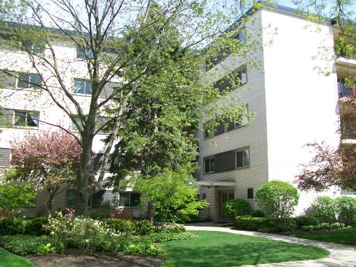Front view of a multi-story brick apartment building with trees and shrubs in the foreground.