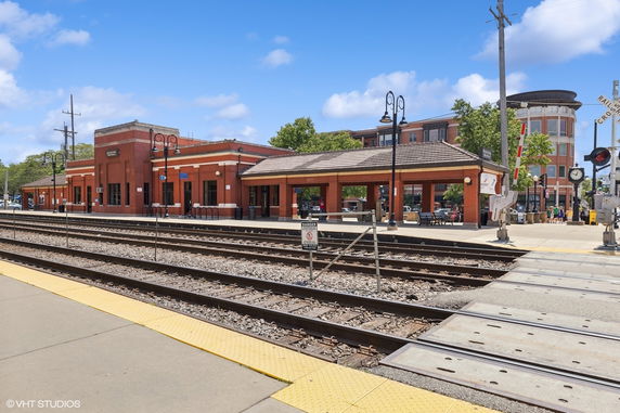 Front view of a red brick railway station with multiple tracks and a crossing barrier.