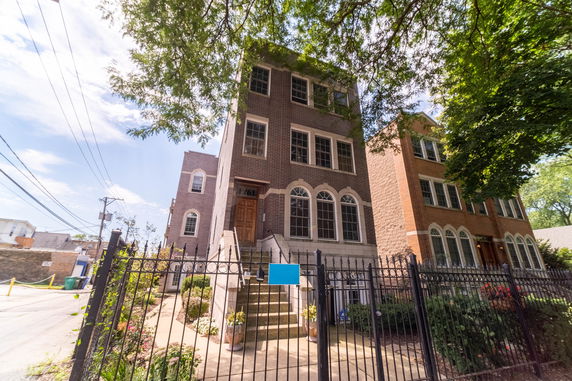 Front view of a three-story brick building with large windows and a fenced entrance.