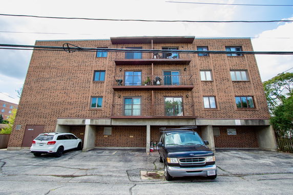 Front view of a multi-story brick building with balconies and ground floor parking spaces.