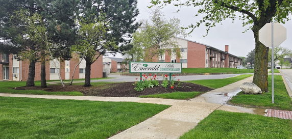 Front view of a condominium building with a sign labeled 'Emerald Hills Condominiums' and a well-maintained lawn.