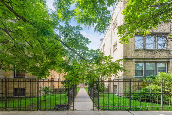 Front view of a multi-story brick apartment building with a walkway and metal fence.