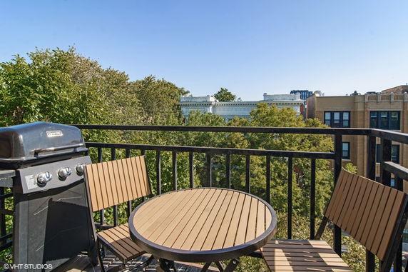 Balcony with chairs and a grill overlooking trees and city buildings.