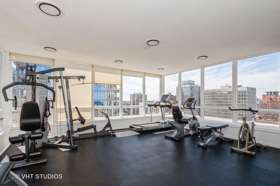 Indoor gym with exercise equipment and large windows overlooking city buildings.