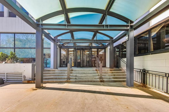 Front view of a building entrance with a glass canopy and stairs leading up to double glass doors.