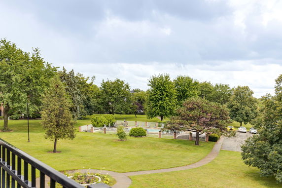 View of a grassy area with trees, a path, parked cars, and a fenced pool area.