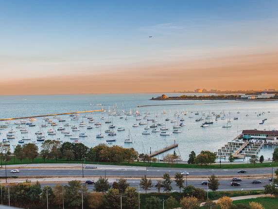 Aerial view of a marina with numerous boats docked in a large body of water, alongside a highway and greenery.