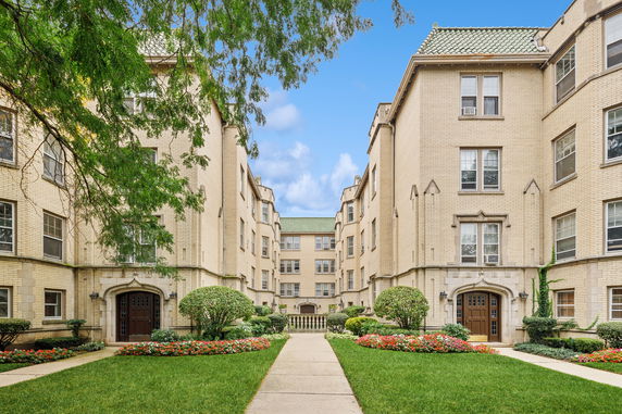 Front view of a multi-story brick apartment building with green tiled roof and landscaped entrance.