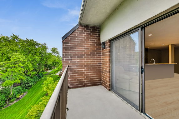 Balcony view from a building with a glass sliding door and a brick wall, overlooking green trees and grass.