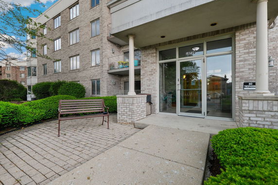 Front view of a multi-story brick apartment building with a glass entrance and a small bench outside.