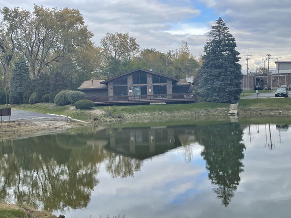 Front view of a single-story building with a sloped roof and large windows, situated next to a pond.