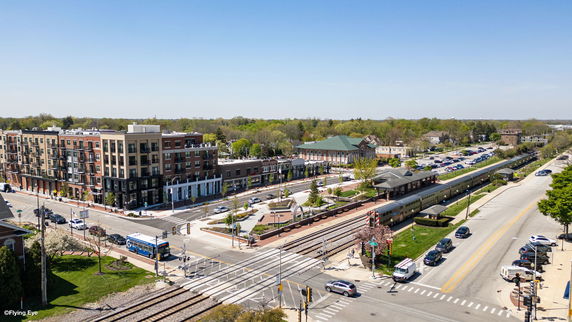 Aerial view of a mixed-use building with multiple floors and nearby train tracks, roads, and green spaces.