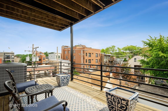 Panoramic view from a rooftop with outdoor seating and railing, overlooking urban buildings and greenery.