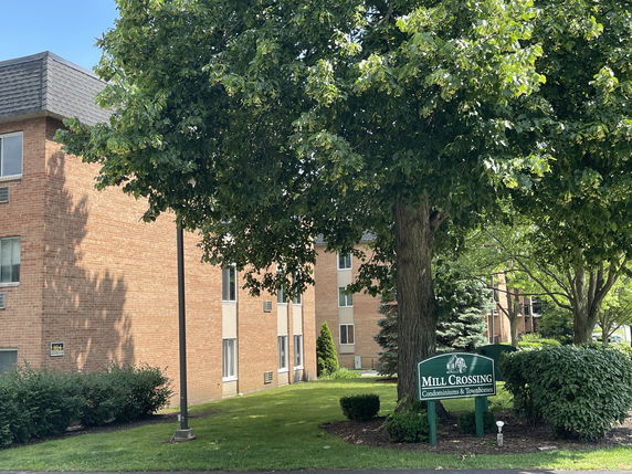 View of a brick condominium building with multiple floors and a green sign reading 'Mill Crossing Condominiums & Townhomes'.