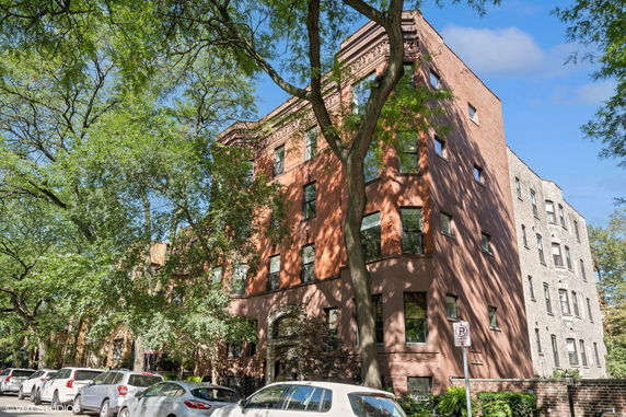 Front view of a multi-story brick building with large trees and parked cars on the street.
