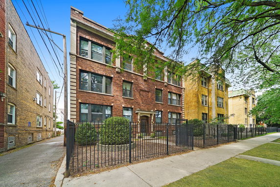 Front view of a multi-story brick apartment building with large windows and a fenced entrance.