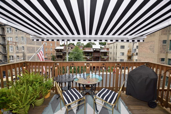Rooftop with seating area, grill, and striped awning overlooking residential buildings.