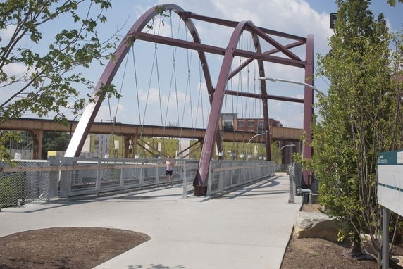 Pedestrian bridge with red arches and railings, surrounded by trees and pathways.