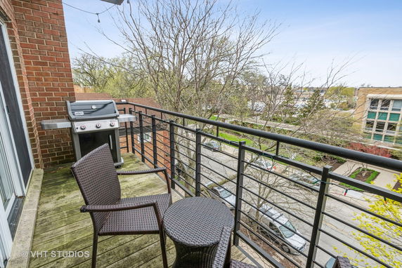 Balcony with chairs, a table, and a grill overlooking trees and a street.