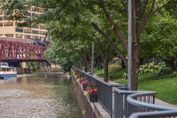 View of a riverwalk with trees, flowers, and a red bridge over a waterway.