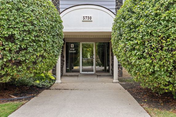 Front view of a building entrance with glass doors and tall bushes on either side.
