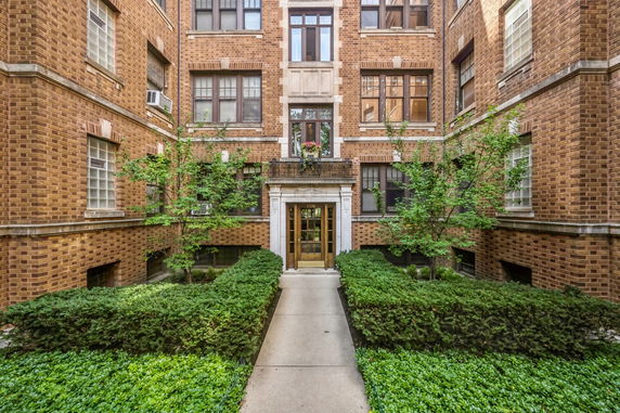 Front view of a brick apartment building with multiple windows and an entrance surrounded by greenery.