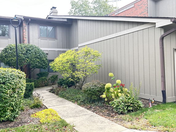 Front view of a two-story house with a sloped roof and chimney, surrounded by a garden.