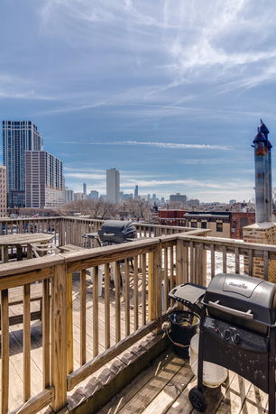 Rooftop area with wooden railing and barbecue grills, overlooking a city skyline.