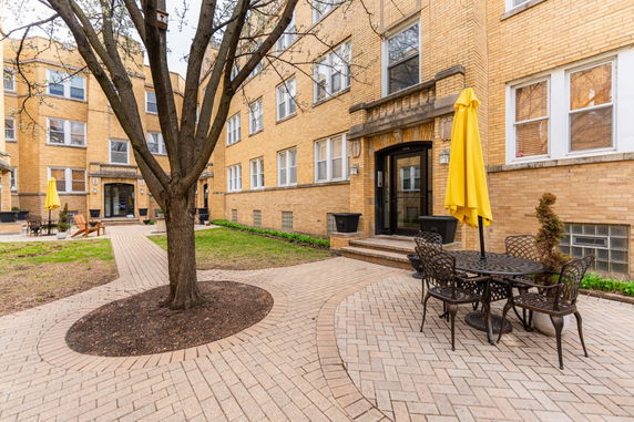Front view of a multi-story brick apartment building with outdoor seating and a tree.