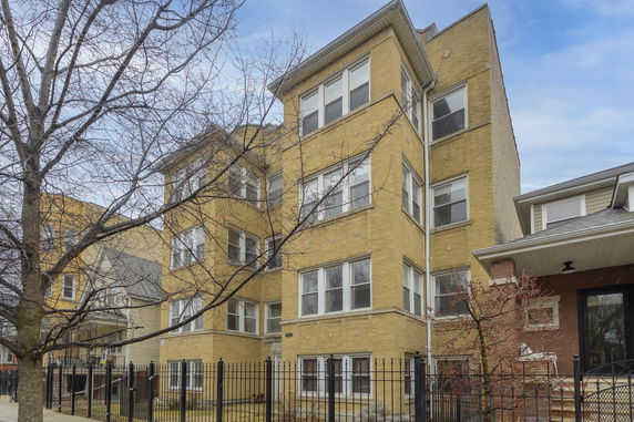 Front view of a four-story yellow brick building with multiple windows and a black metal fence.