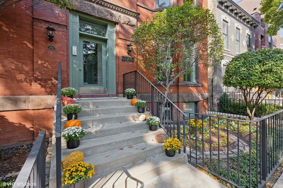 Front view of a brick building with a green door and steps decorated with plants and pumpkins.