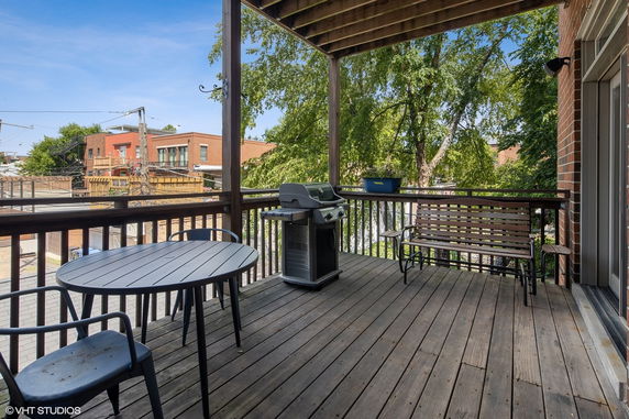Outdoor patio with a wooden deck, table, chairs, grill, and view of nearby buildings and trees.
